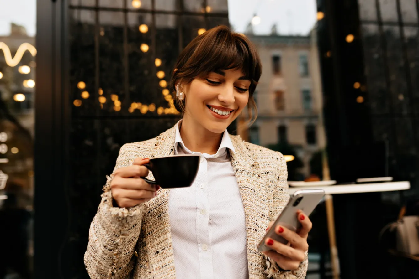 A woman holding a cup of coffee looks at her smartphone screen